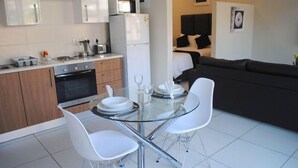 A modern kitchen with white cabinets, a refrigerator, and a microwave. There is a dining area with a glass table and white chairs. - 86 Edison Guest Lodge (Sandton)