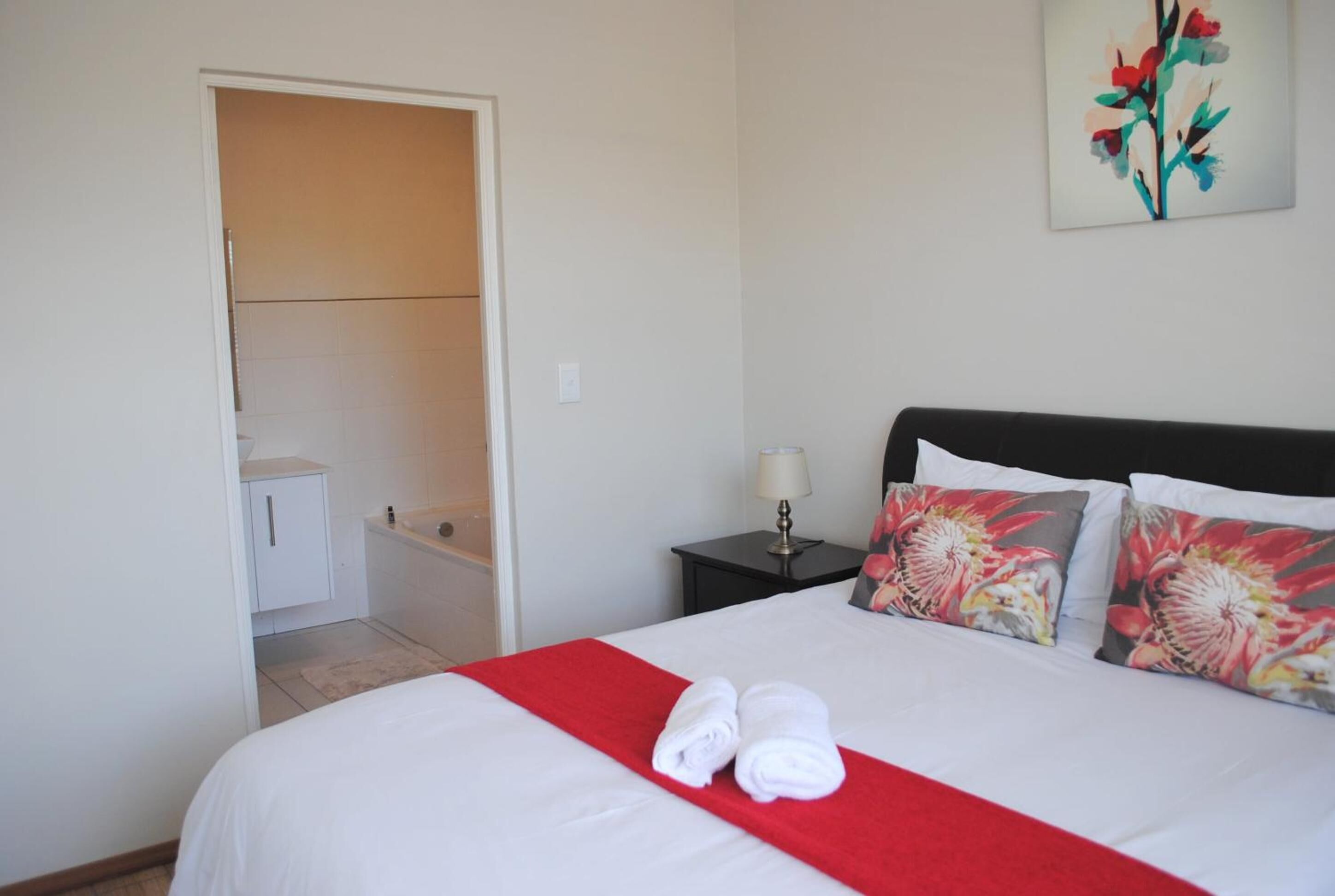 A neatly made bed with white bedding and a red accent, a nightstand with a lamp, and a painting on the wall.