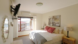 Et soveværelse med seng, et vindue med gardiner, en vægmonteret aircondition, et dekorativt spejl, en lampe og et lille trænatbord.