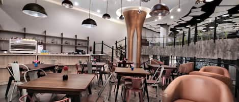 Un interior de restaurante moderno con varias mesas y sillas, una columna central y lámparas colgantes.
