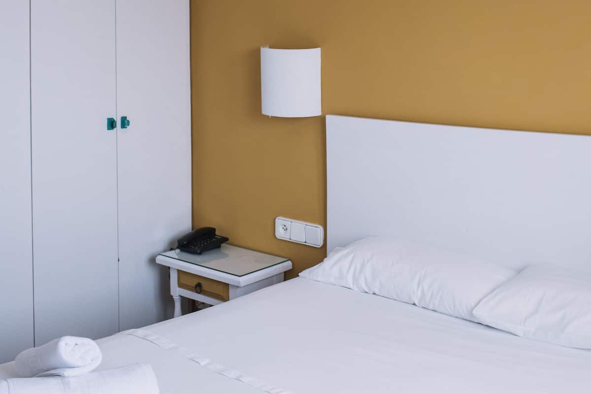 A hotel room with a bed, white linens, a nightstand with a phone, and a wardrobe.
