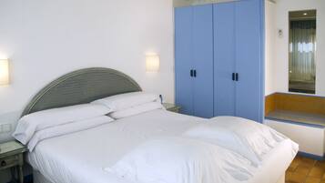 Una cama con sábanas blancas, una mesita de noche, un armario azul y una ventana pequeña con vistas.