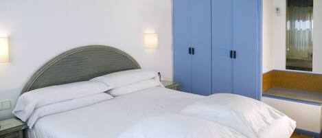 Una cama con sábanas blancas, una mesita de noche, un armario azul y una ventana pequeña con vistas.