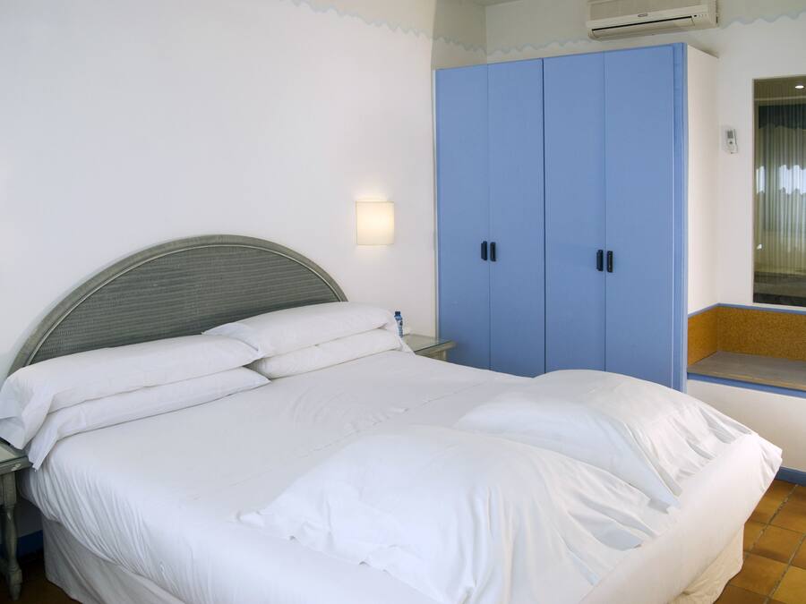 Una cama con sábanas blancas, una mesita de noche, un armario azul y una ventana pequeña con vistas.
