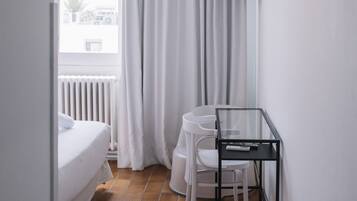 A small, modern room with a bed, a desk with a chair, a TV mounted on the wall, and a window with curtains.