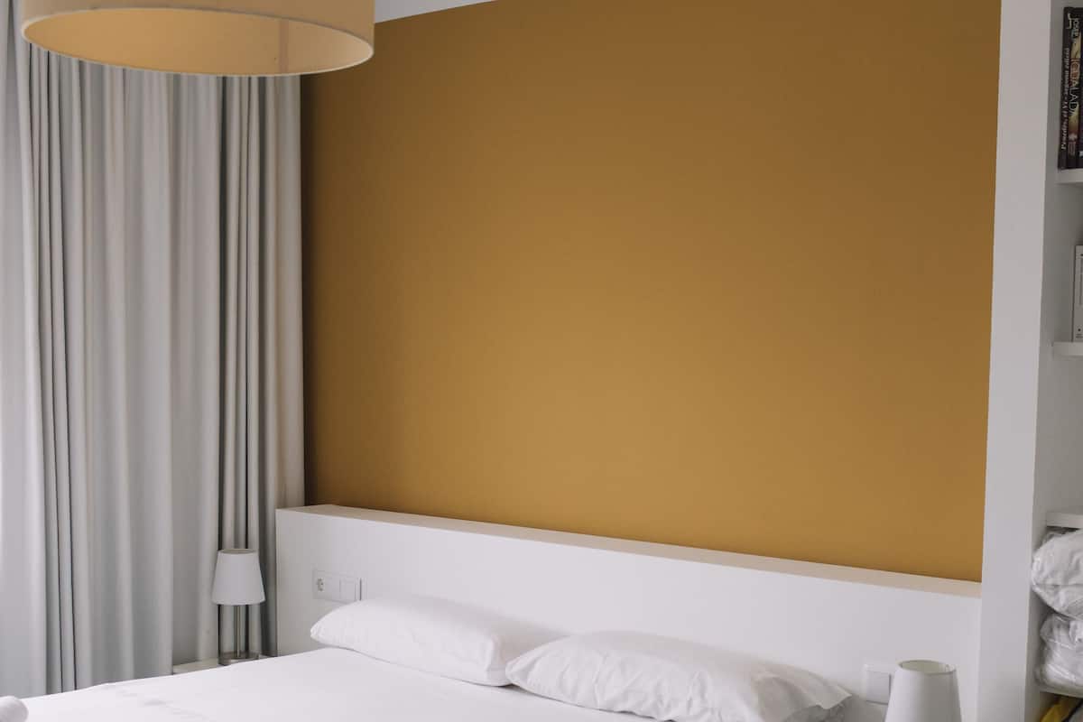 A modern hotel room with a large bed, white bedding, a beige headboard, a bedside table with a lamp, and a curtain.