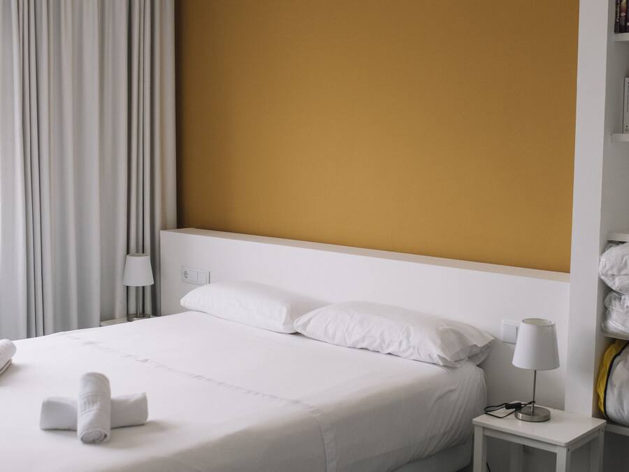 Una habitación de hotel moderna con una cama grande, ropa de cama blanca, un cabecero color beige, una mesita de noche con una lámpara y una cortina.
