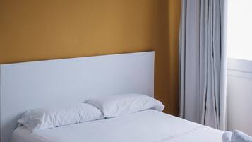 A neatly made bed with white linens and towels, a white headboard, and a window with curtains.