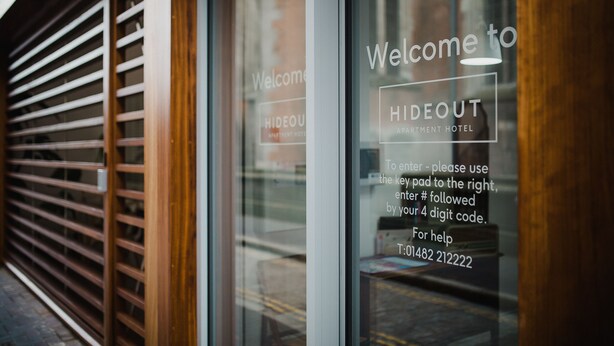 A hotel entrance with a welcome sign, a keypad for entry, and a help phone number. - Hideout Apartment Hotel (Hull)