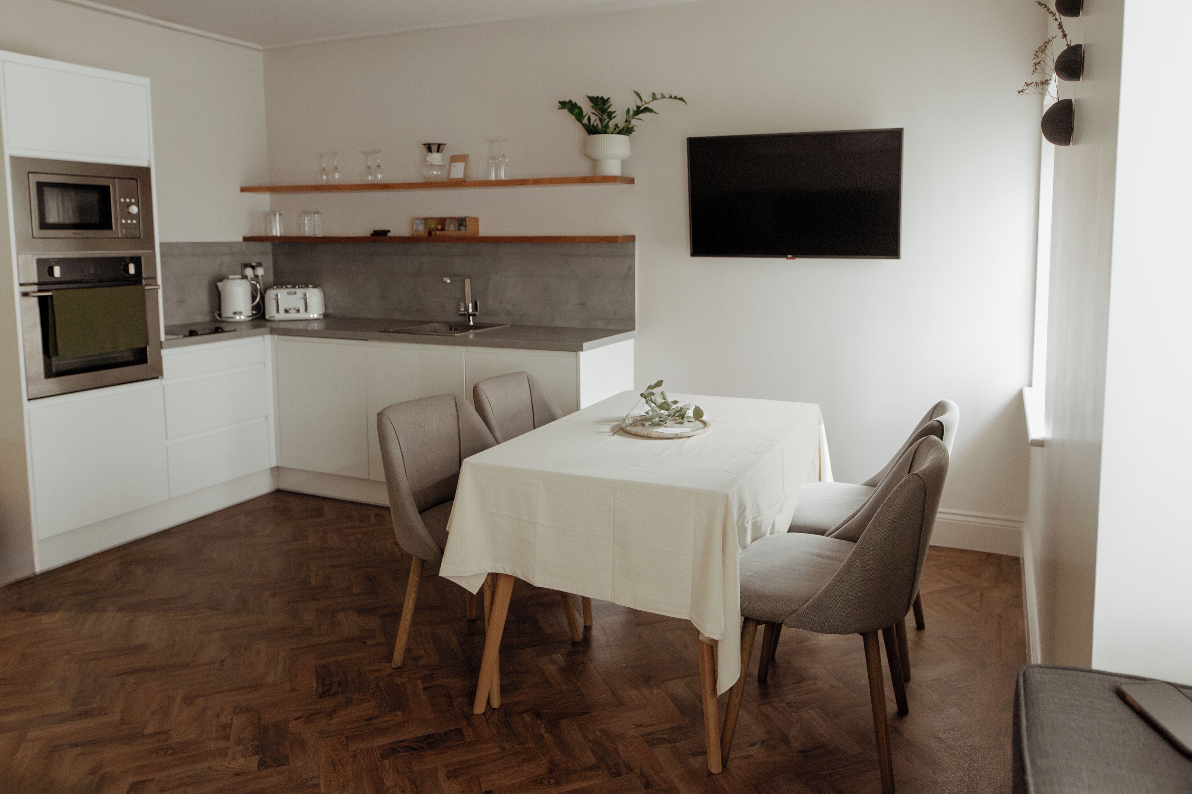 A modern kitchen with white cabinets, a built-in oven, and a microwave. There is a dining table with chairs and a potted plant on the wall.