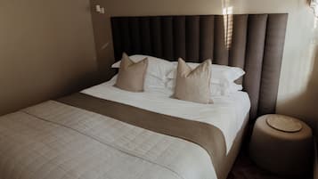 Una cama bien hecha con cabecera, un otoman redondo y una lámpara de pared.