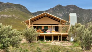 A wooden cabin with a balcony, surrounded by mountains and sparse vegetation. - AfriCamps at Stanford Hills Estate- Glamping (Stanford)