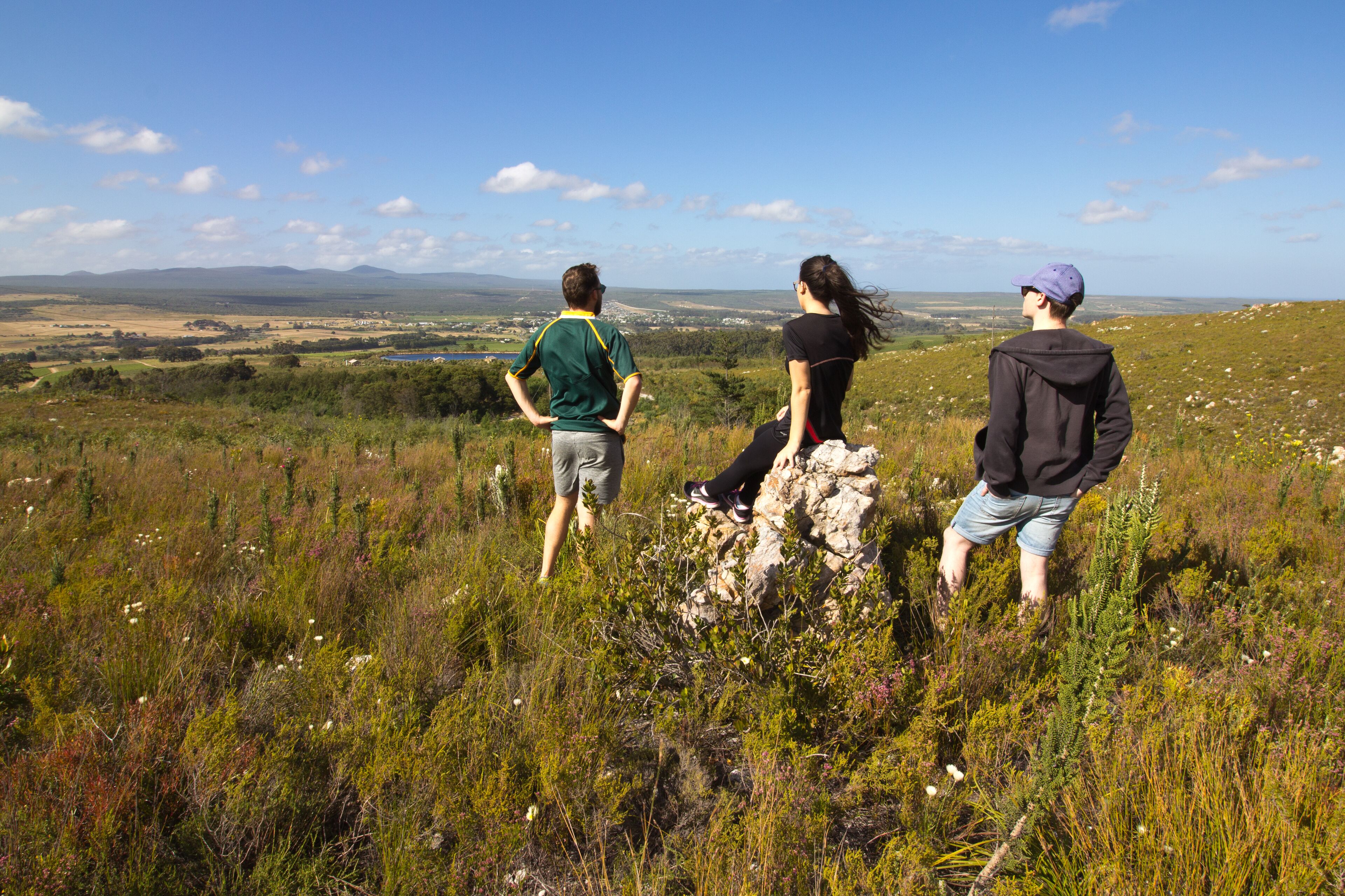 Three people standing on a grassy hill overlooking a landscape with distant mountains and a clear sky.