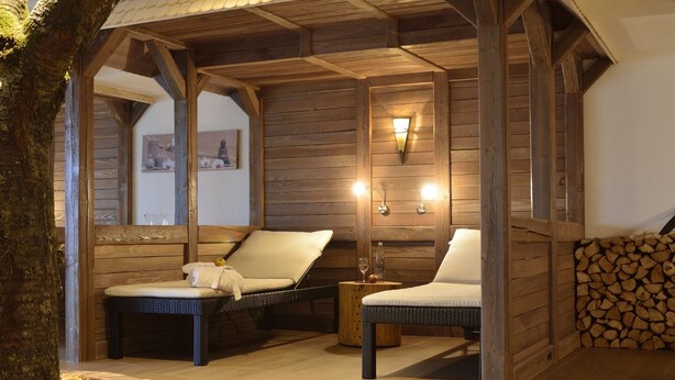 A cozy wooden cabin with two wicker chairs, a small table with a glass, and a wall-mounted light fixture. - Burg Hotel (Feldberg)
