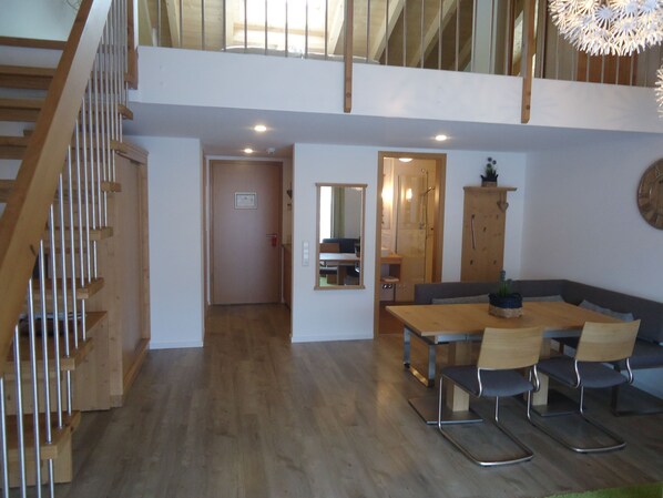 Amplia sala de estar con escalera de madera, una mesa de comedor con sillas, un sofá y una cocina visible a través de un umbral abierto.