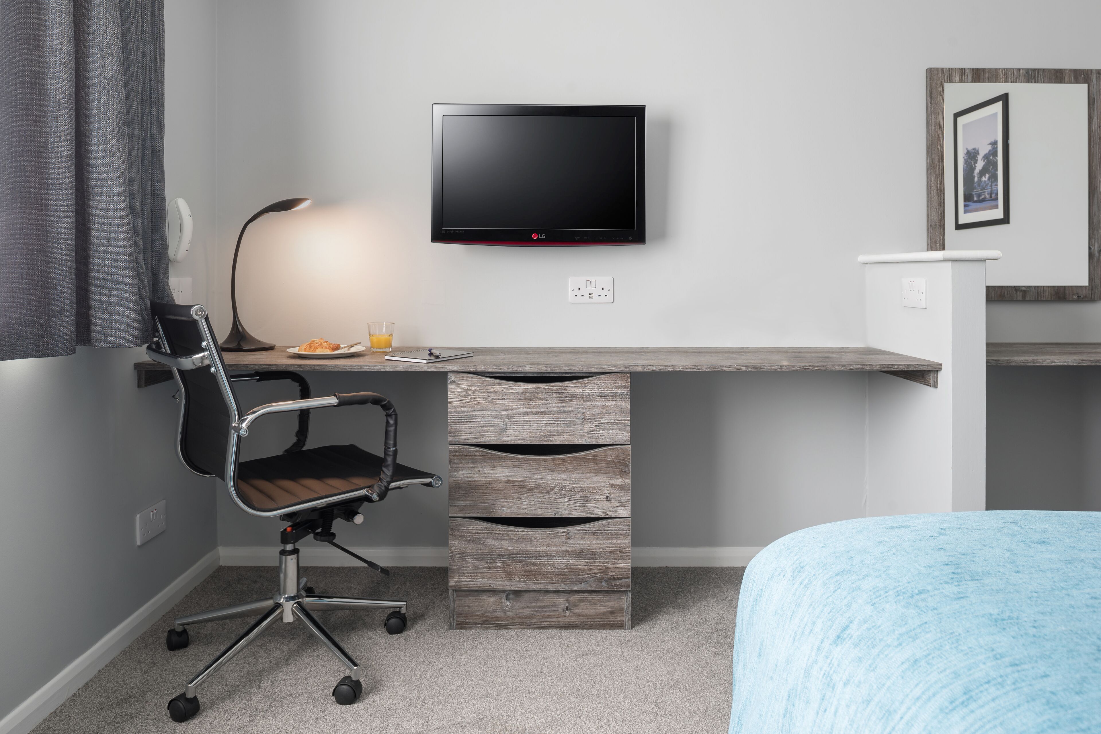 Un dormitorio moderno con cama, escritorio con monitor de computadora, silla y mesita de noche.