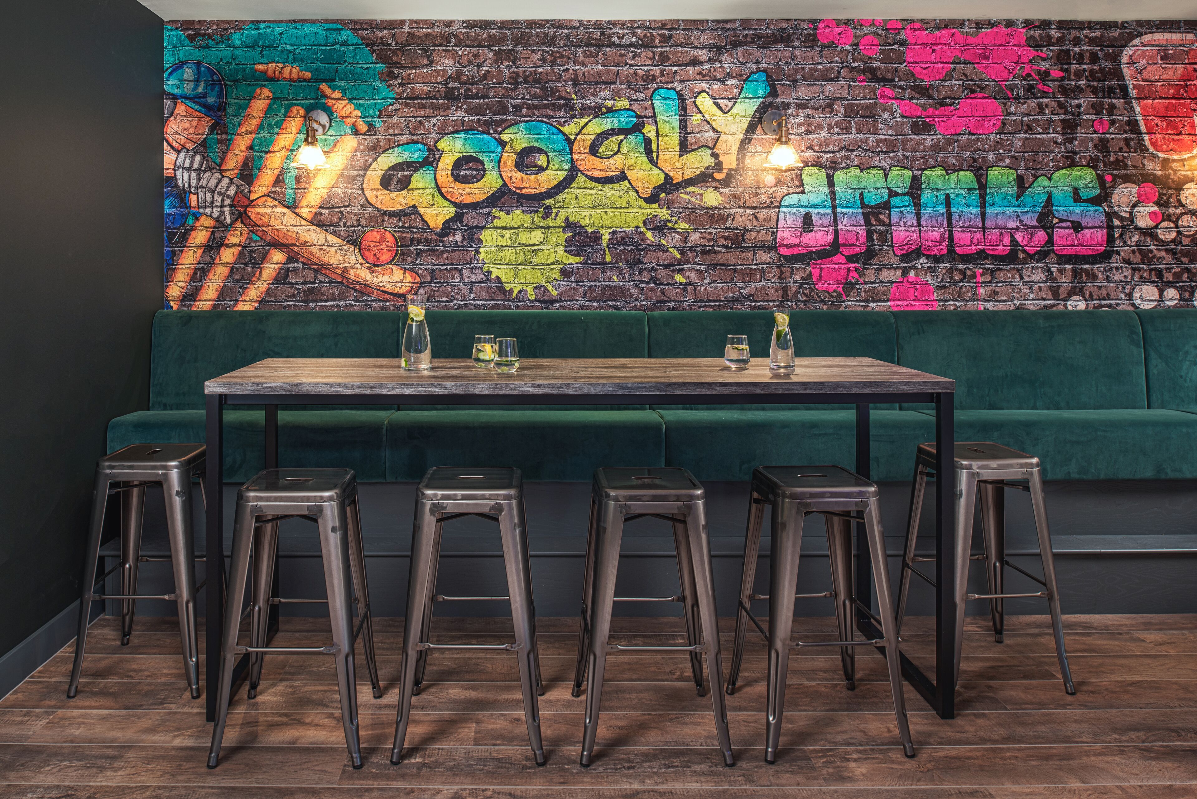 Zona de bar con un muro lleno de grafitis, un banco acolchado verde y taburetes de metal.