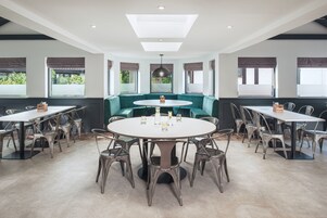 A modern dining area with a round table surrounded by metal chairs, a green sofa, and large windows with blinds.