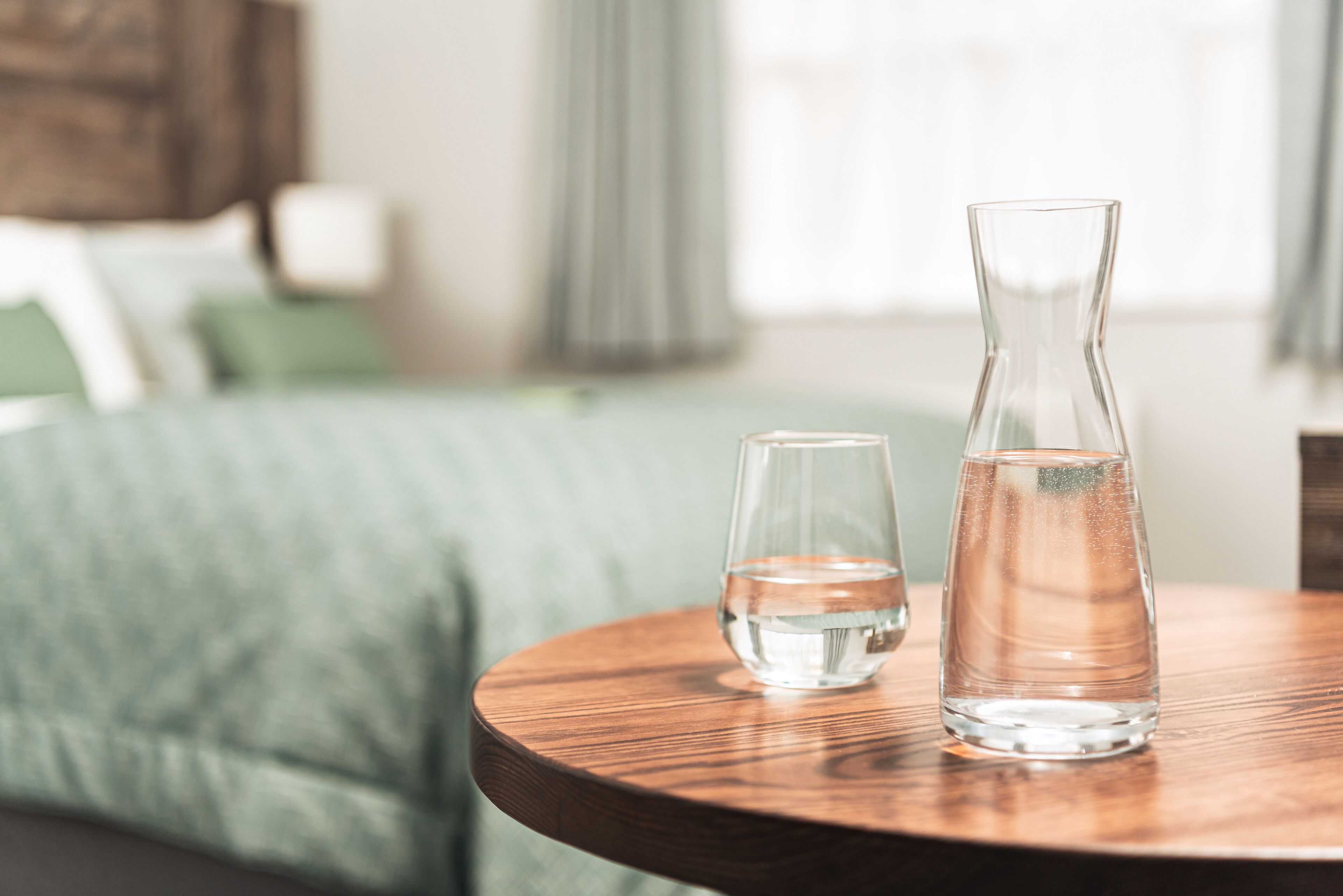 Een houten tafel met twee heldere glazen flessen, één hoger en één lager, in een kamer met een groen gestoffeerde bank en een raam met gordijnen.