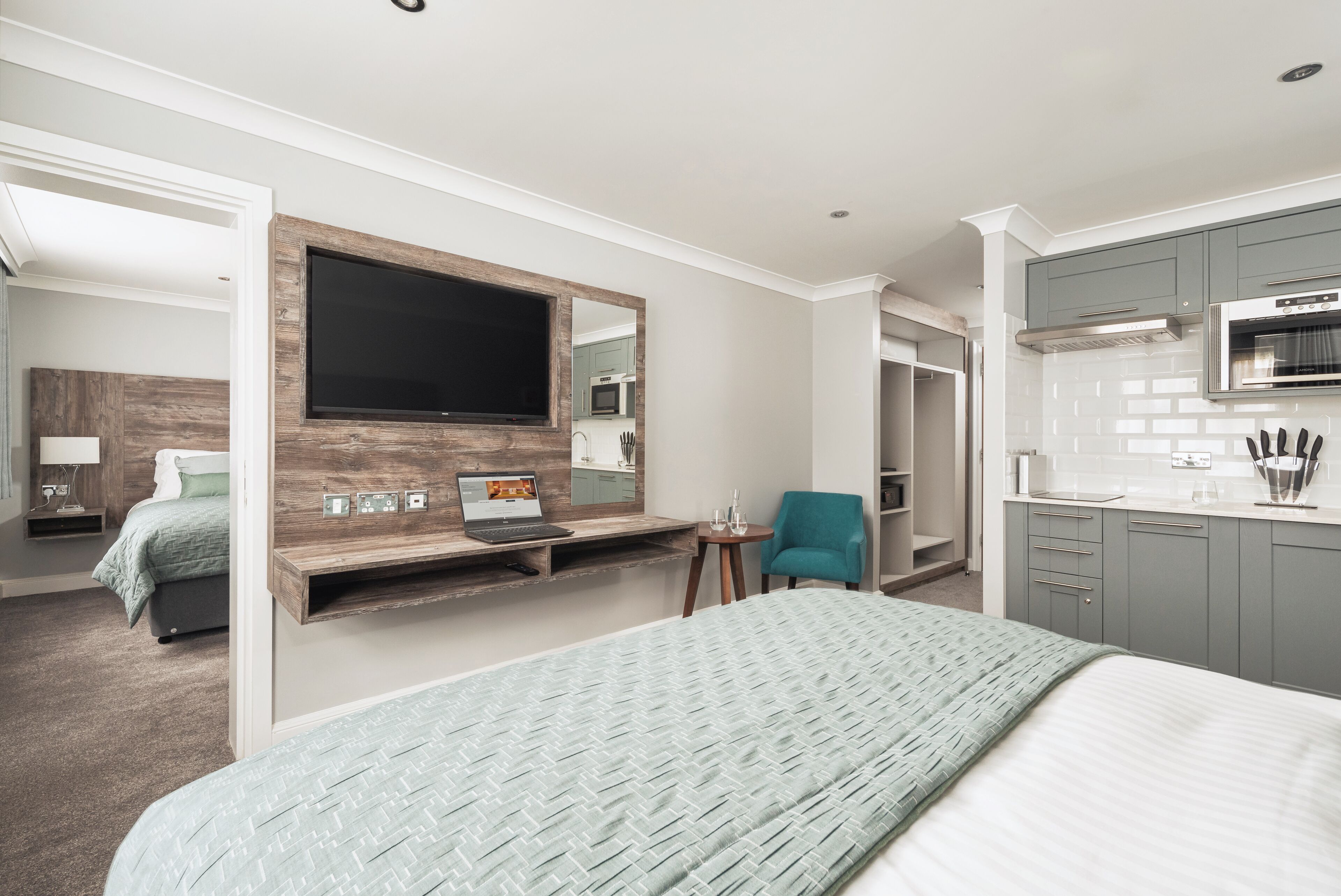 Una habitación de hotel moderna con una cama grande, un escritorio con una computadora portátil, un televisor montado en un panel de madera y una kitchenette con armarios grises.