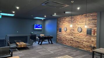 A modern lounge area with a foosball table, comfortable seating, a TV screen, and a dartboard on a wooden wall.