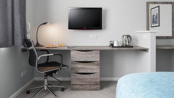 Una habitación de hotel moderna con una televisión de pantalla plana montada en la pared, un escritorio con lámpara, una silla y una cama con manta azul.