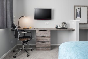 Una habitación de hotel moderna con una televisión de pantalla plana montada en la pared, un escritorio con lámpara, una silla y una cama con manta azul.