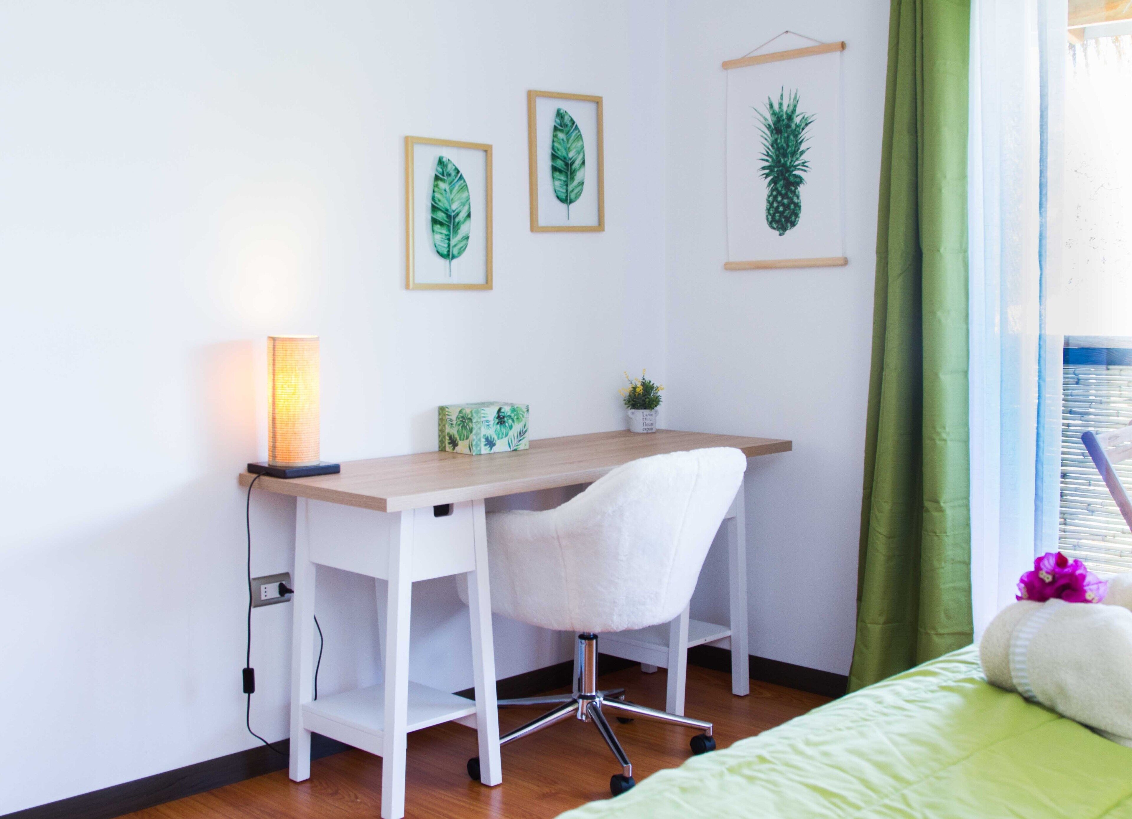 Ein ordentlich eingerichtetes Schlafzimmer mit einem Holztisch, einem weißen Stuhl, einem Bett mit grüner Bettwäsche und einem Fenster mit grünen Vorhängen.