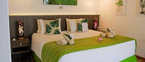 A neatly made bed with a green bedspread, multiple pillows, and a headboard. There is a nightstand with a decorative lamp and a small table with a plant.