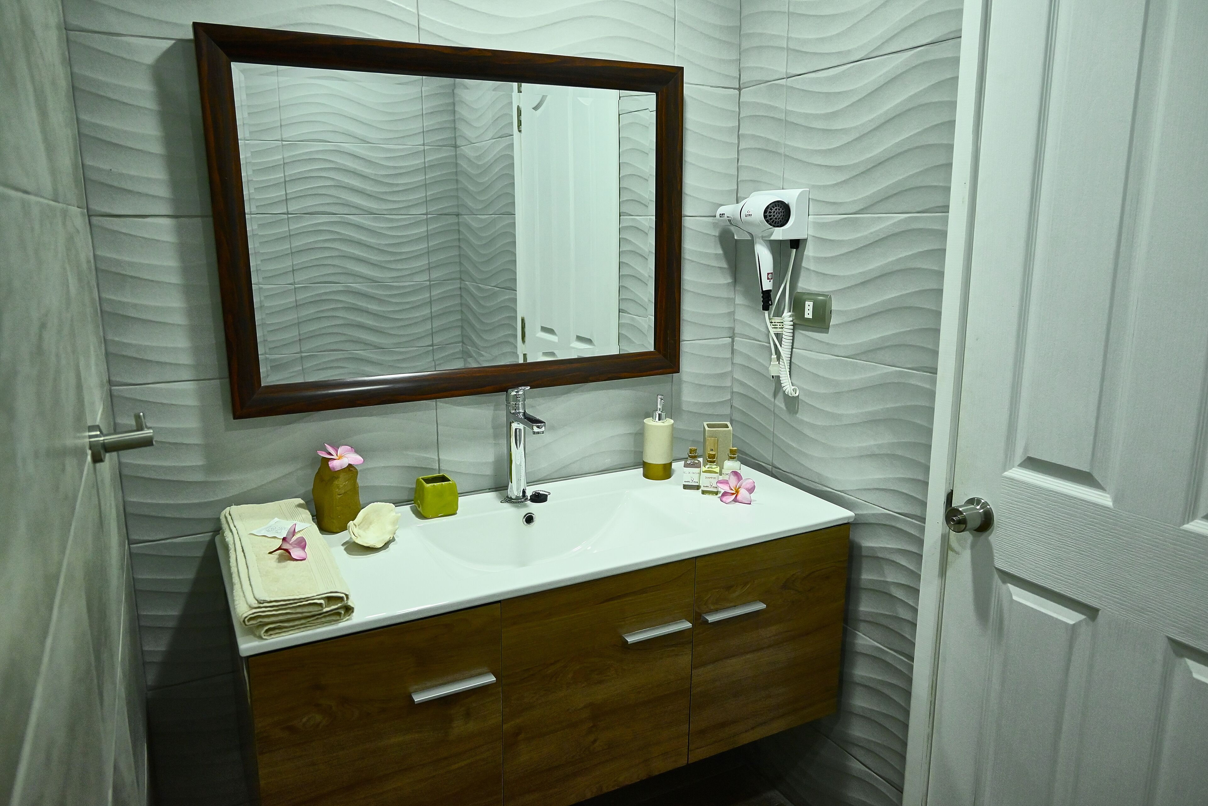 A bathroom with a white sink, wooden vanity, a large mirror, and a hairdryer on the wall.