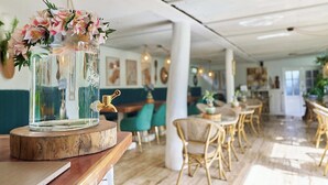 A dining area with wooden tables and chairs, a glass vase with flowers on a wooden stand, and a white ceiling with hanging lights. - Hare Nua (Hanga Roa)