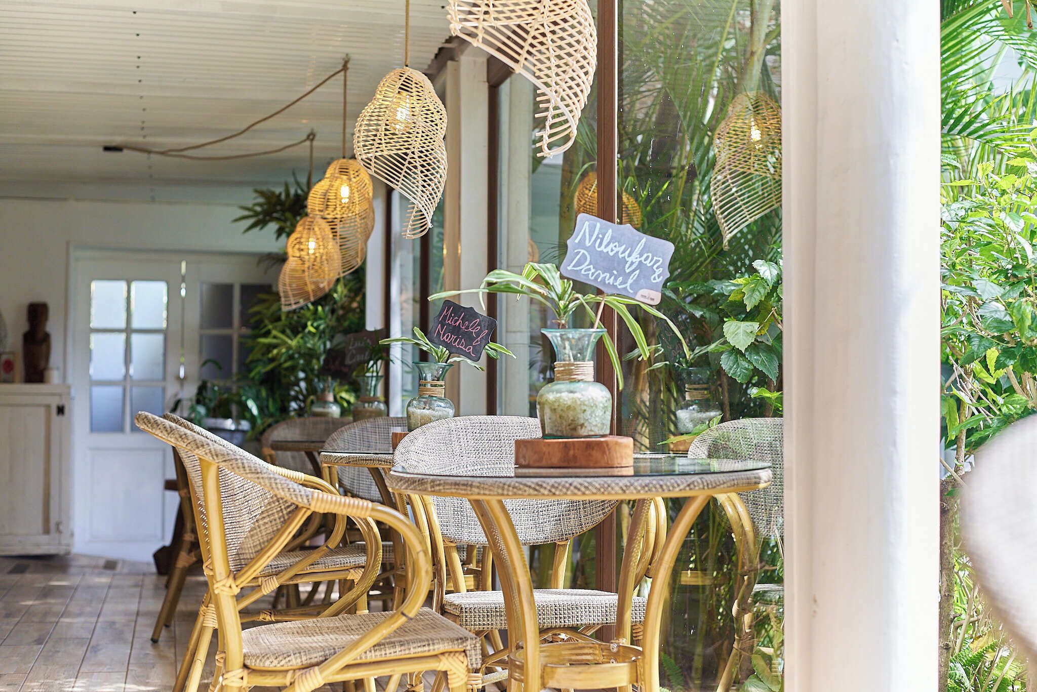 A dining area with wicker chairs and tables, a glass-top table with a vase and a chalkboard sign, and hanging wicker lamps.