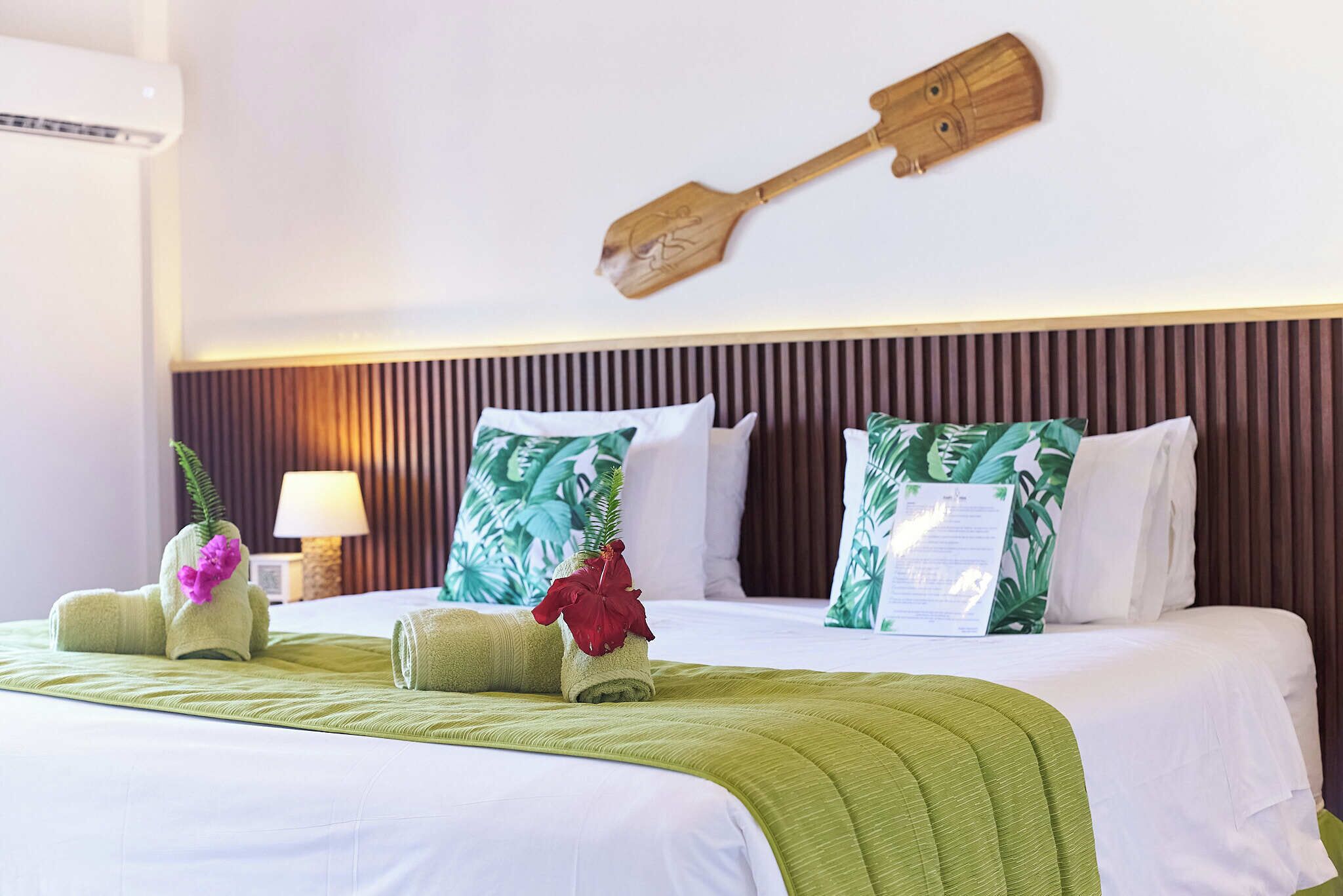 A hotel room with a bed, green towels, a wooden paddle wall decoration, and tropical-themed pillows.