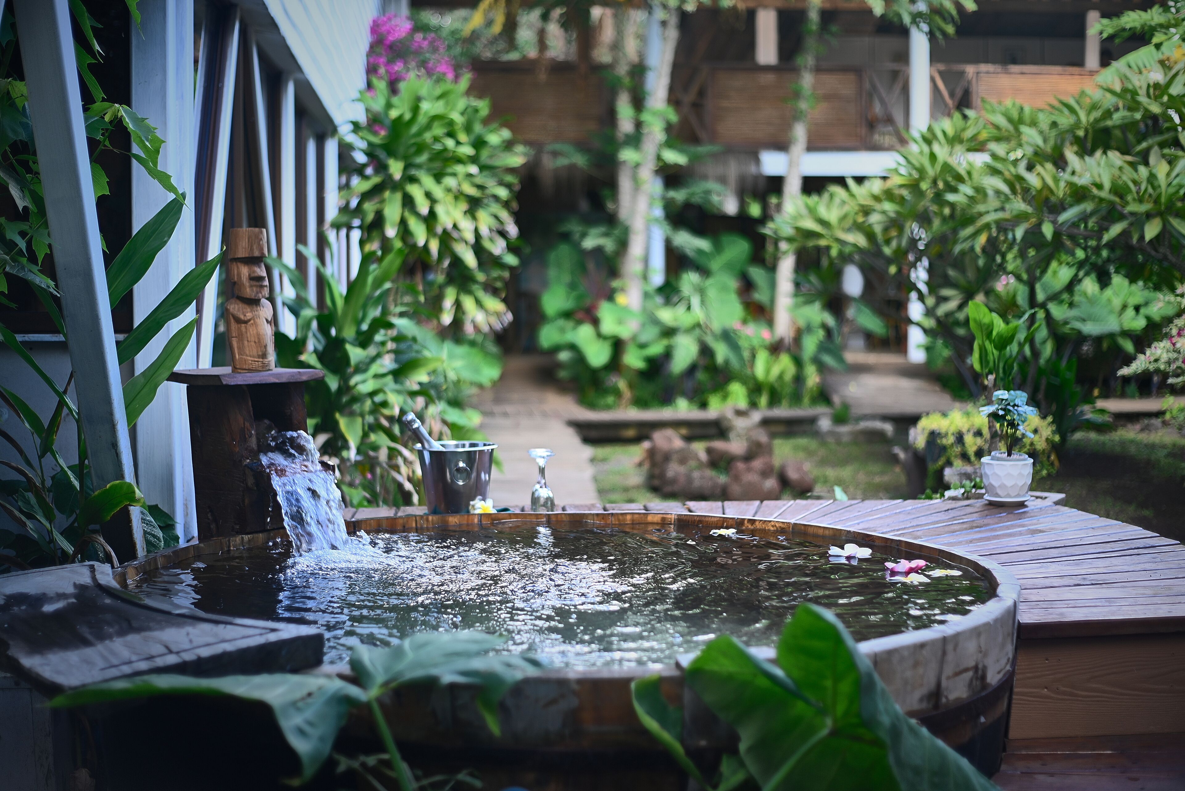 Una terraza de madera con un estanque circular, una estatua de madera y una pequeña cascada.