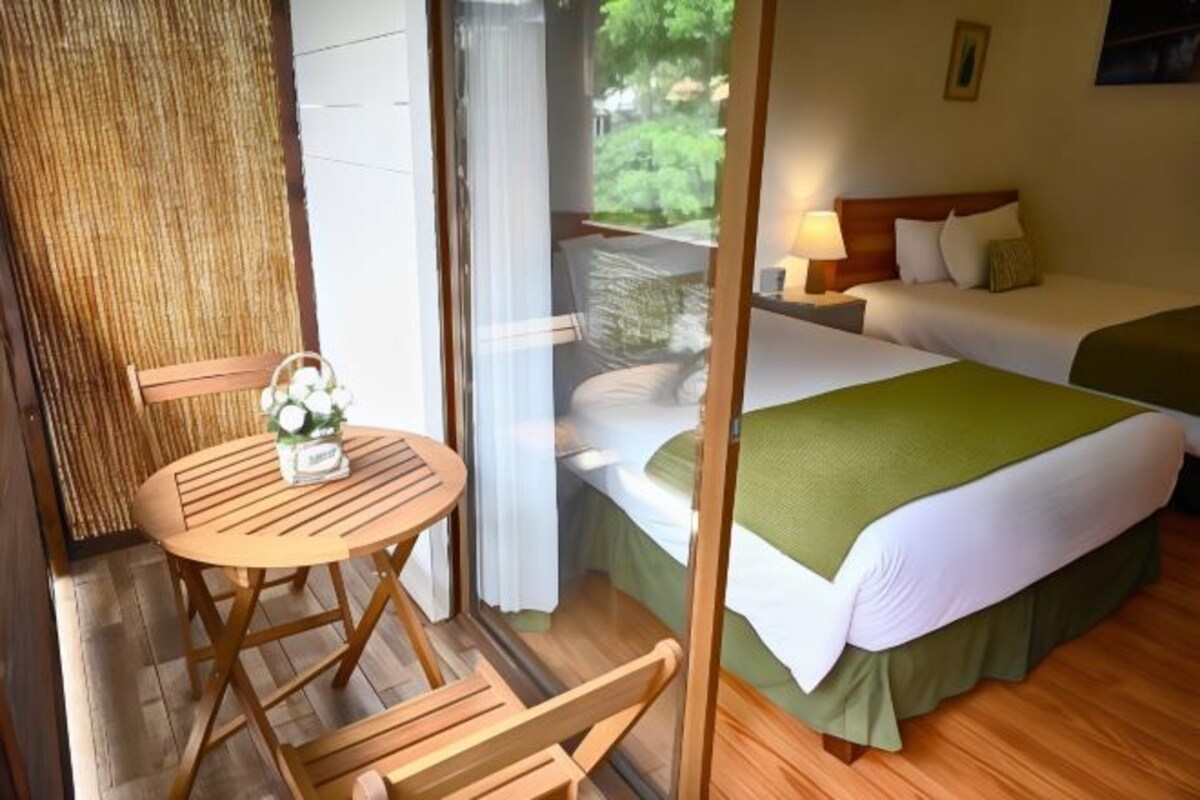 A room with a bed, a small table with chairs, and a view of greenery outside.