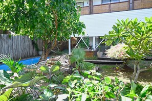 A backyard with a pool, various plants, a wooden deck, and a white building in the background.