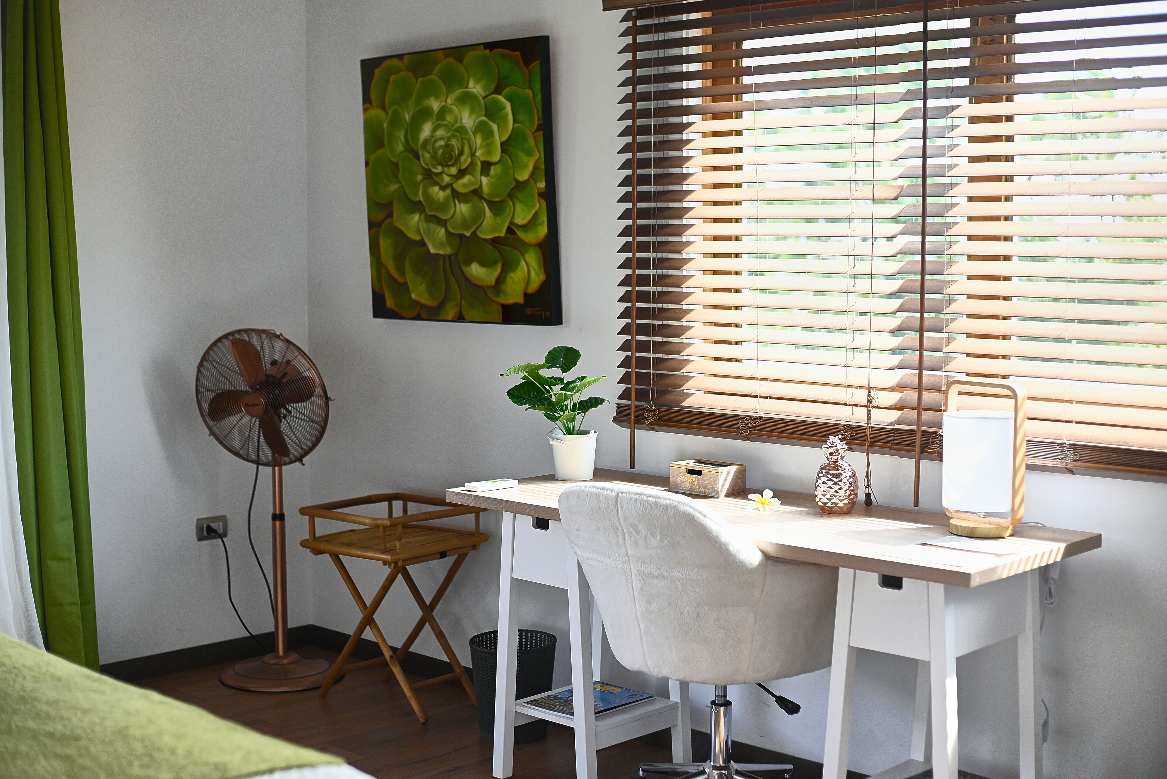 Um quarto com escrivaninha, cadeira, ventoinha, planta em vaso e janela com persianas.