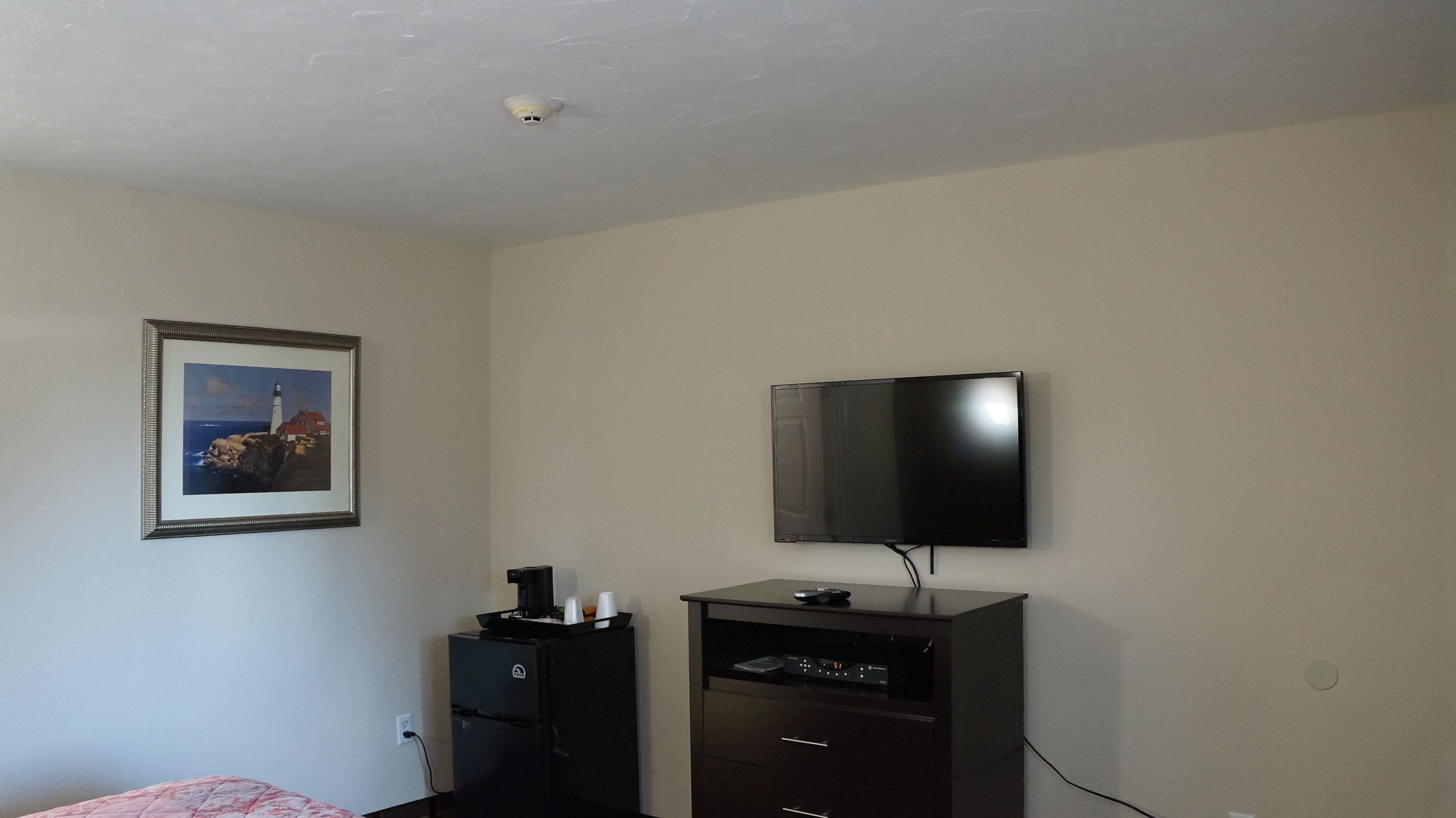 Ein Zimmer mit einem an der Wand montierten Flachbildfernseher, einem kleinen Kühlschrank und einem gerahmten Bild eines Leuchtturms.