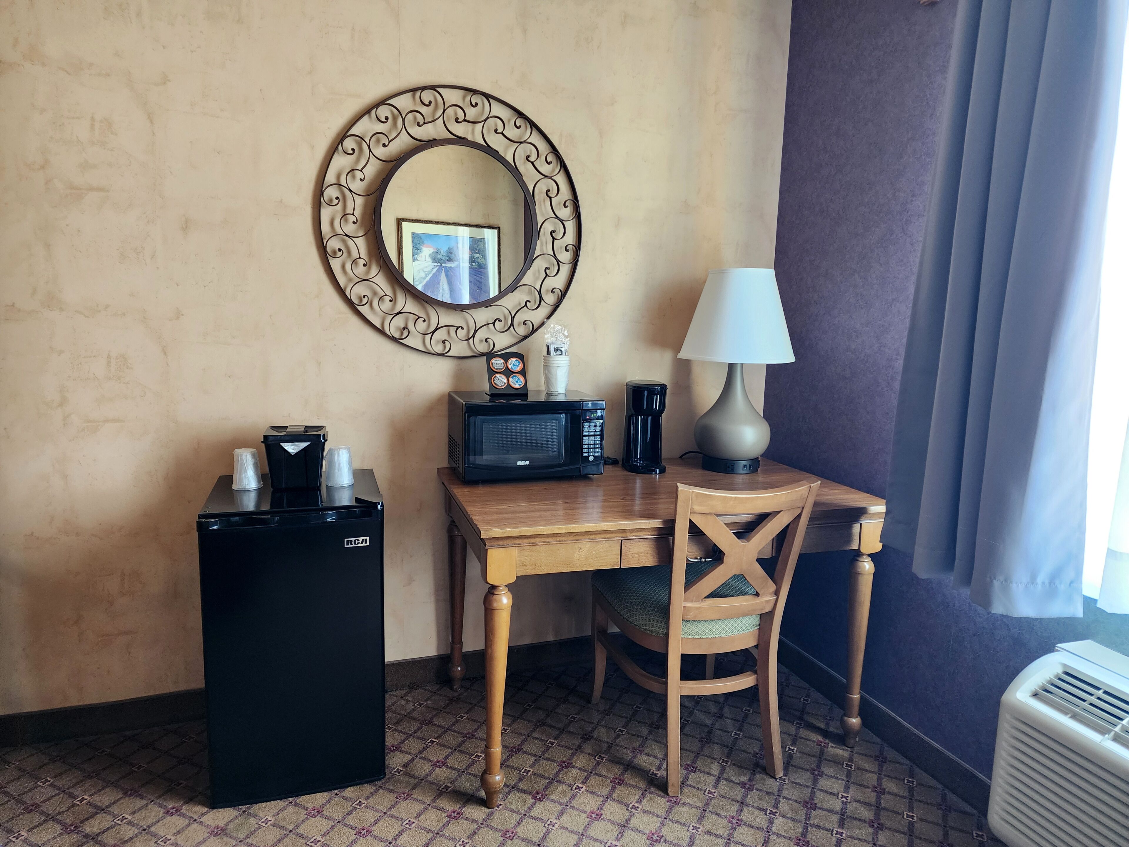 Habitación de hotel pequeña con escritorio de madera, refrigerador negro, microondas, cafetera, lámpara y una silla.