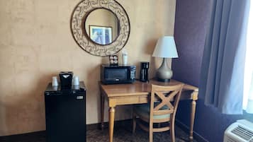 Habitación de hotel pequeña con escritorio de madera, refrigerador negro, microondas, cafetera, lámpara y una silla.