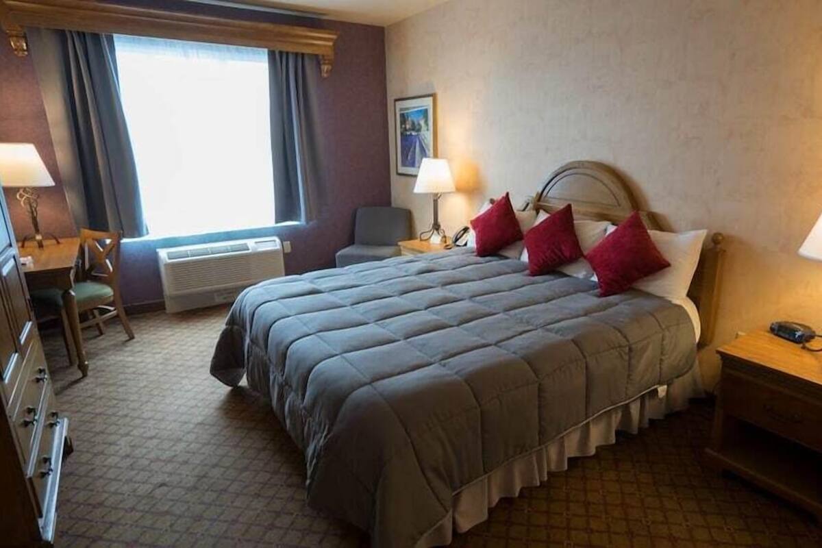 A hotel room with a bed, two bedside tables, a desk, a chair, a window with curtains, and a wall-mounted picture.