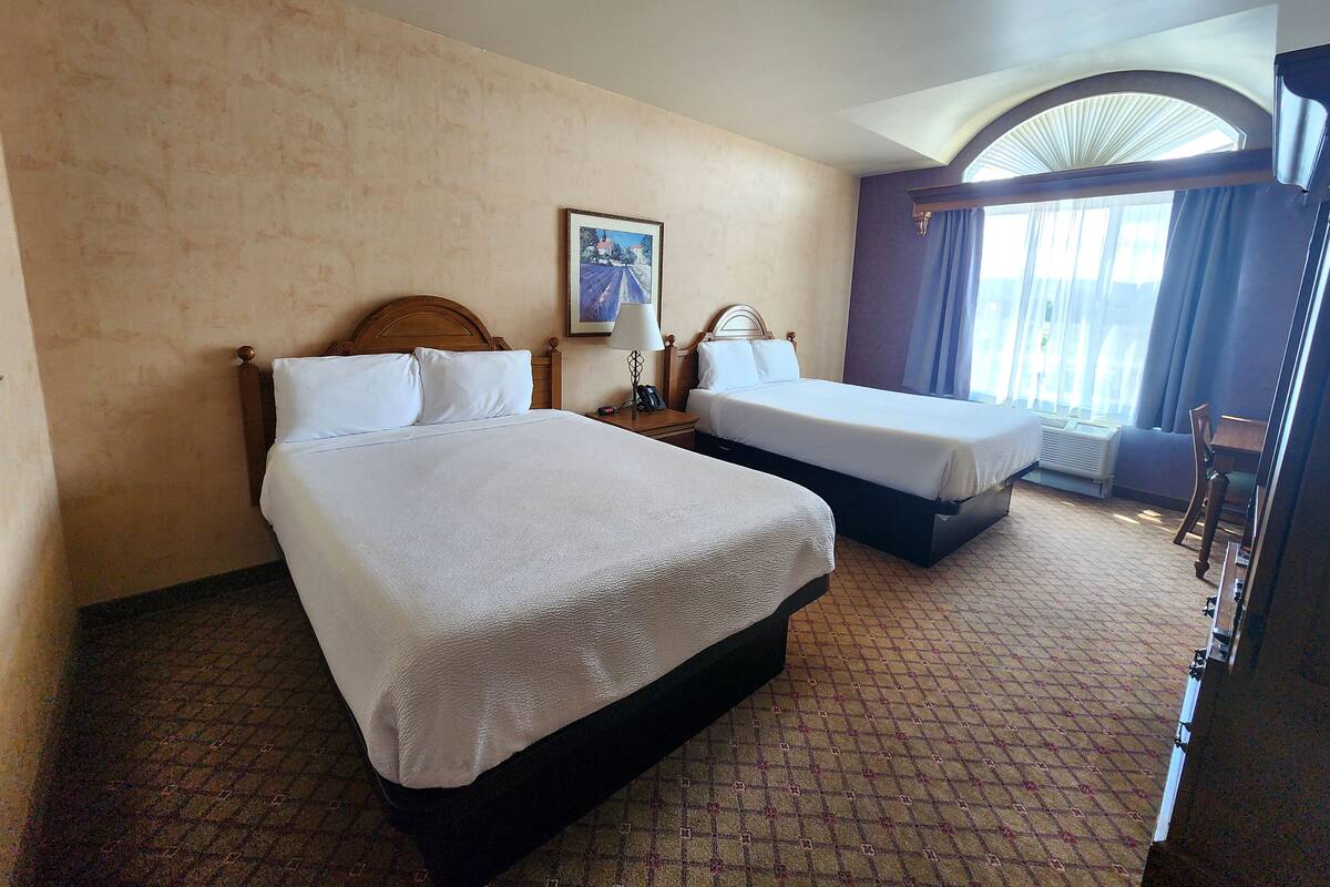 A hotel room with two beds, a painting on the wall, a window with curtains, and a patterned carpet.