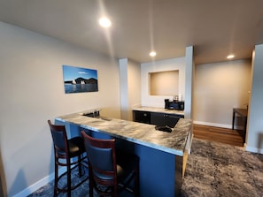 A kitchen with a marble countertop, a sink, a microwave, and a coffee maker. There are two bar stools and a painting on the wall.