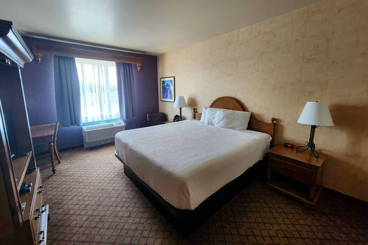 A hotel room with a large bed, a desk, a chair, a window with curtains, a wall-mounted picture, and a bedside lamp.