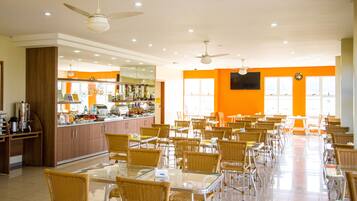 A spacious dining area with multiple tables and chairs, a kitchen in the background, and ceiling fans.