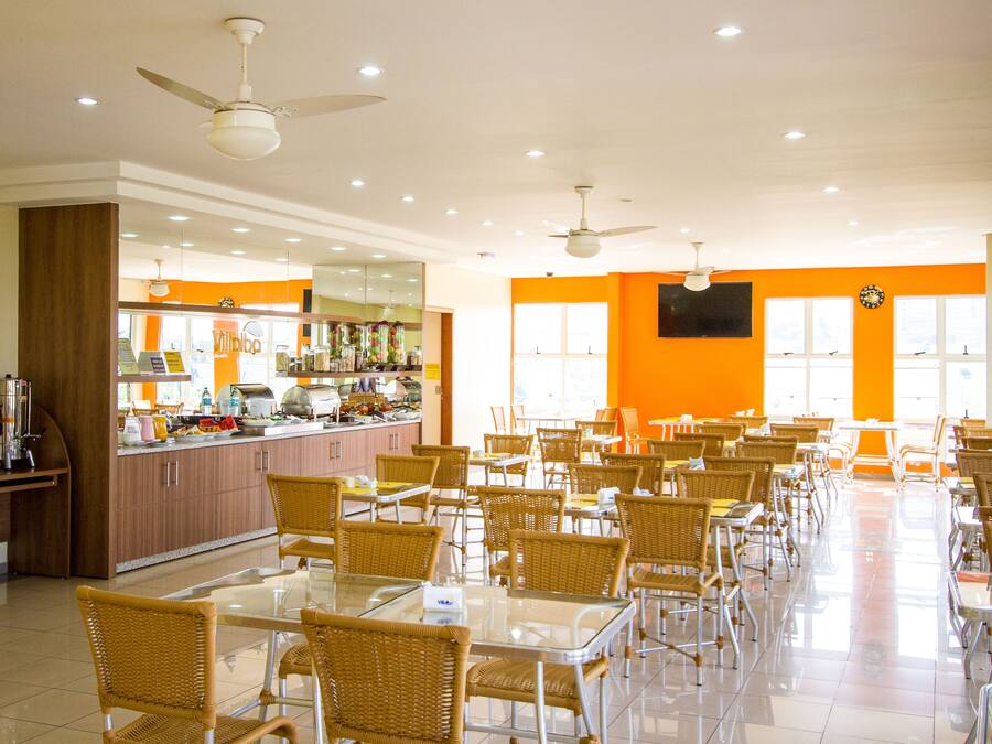 A spacious dining area with multiple tables and chairs, a kitchen in the background, and ceiling fans.