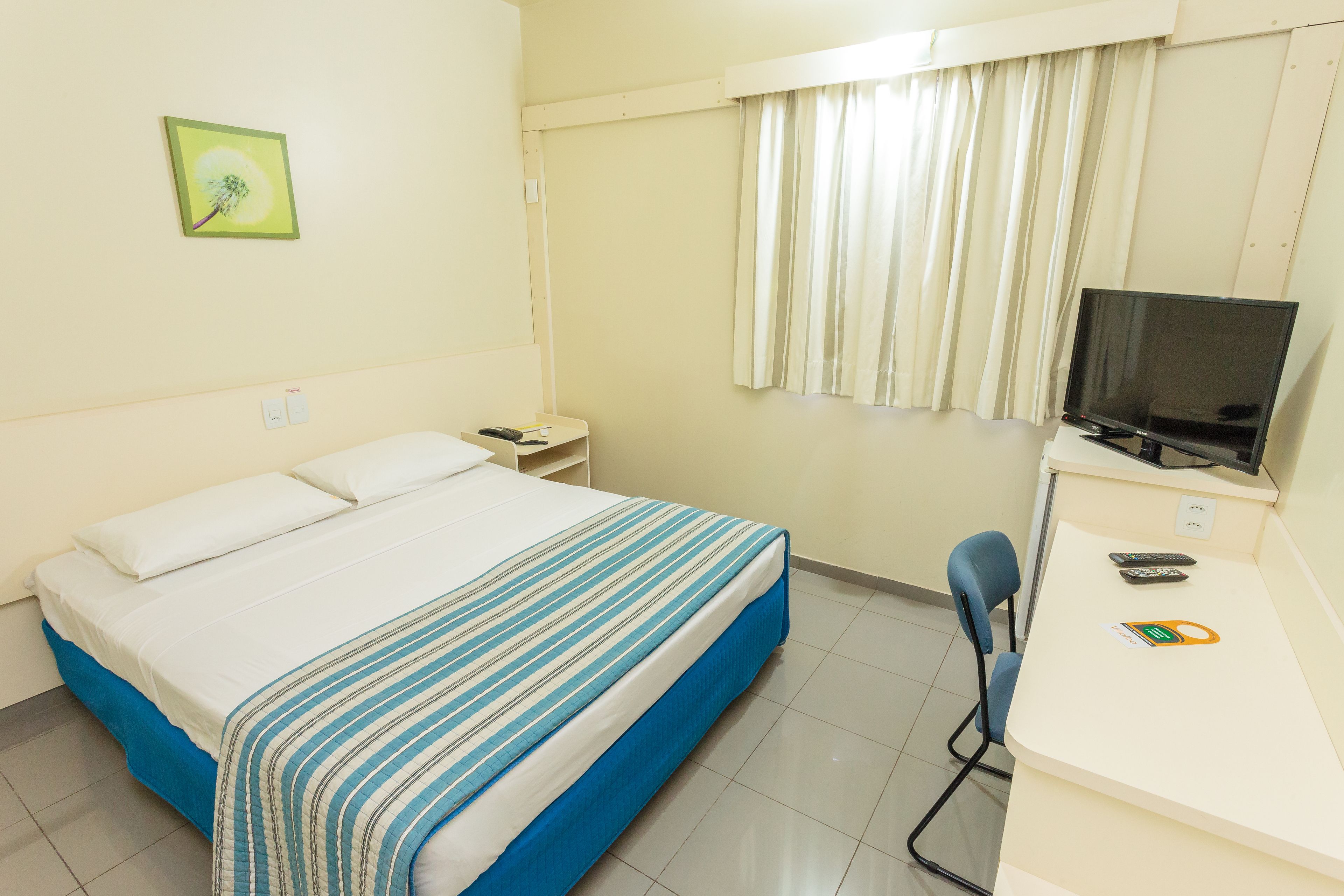 Quarto de hotel com cama, escrivaninha, televisão e cadeira.