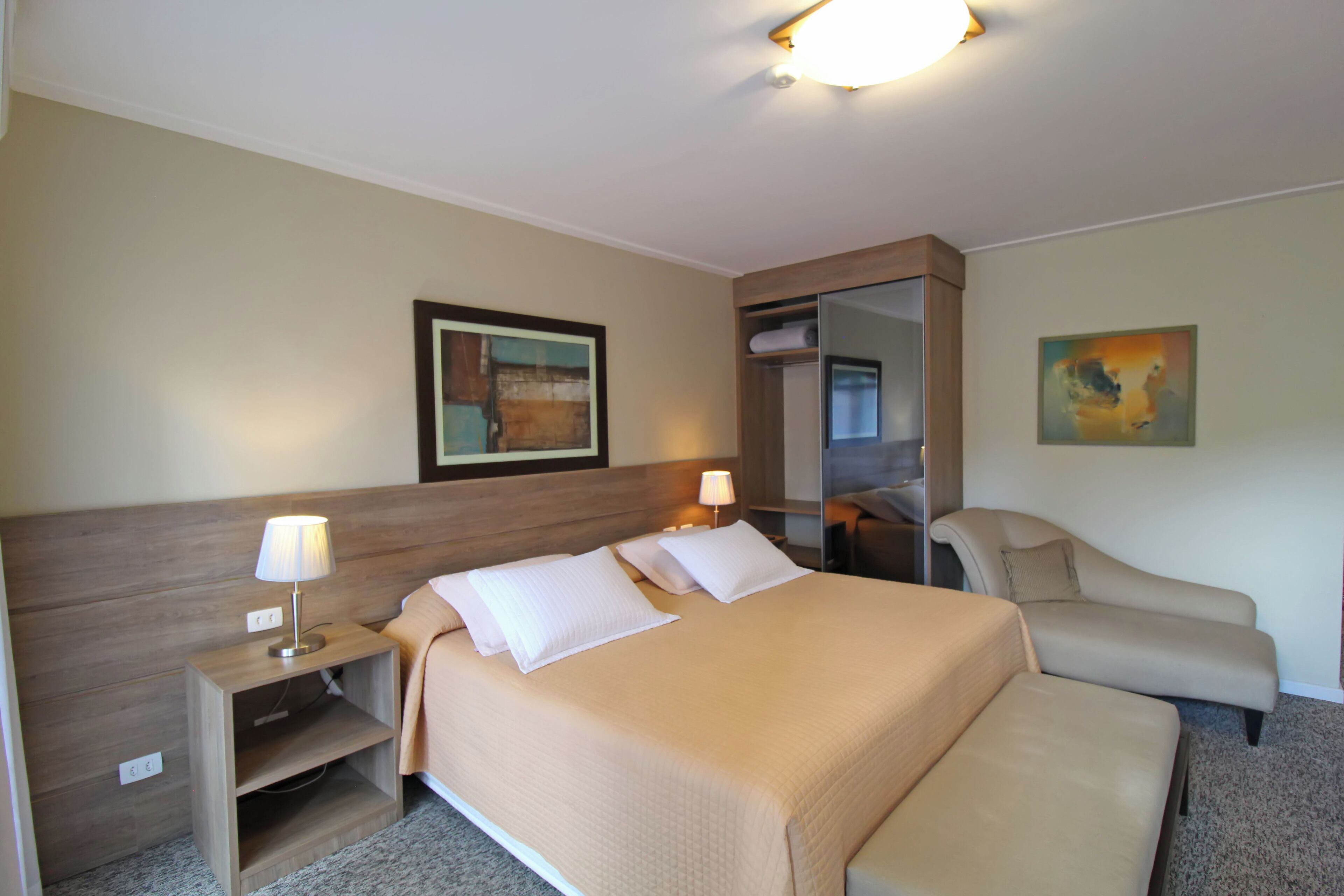 Una habitación de hotel con una cama grande, una mesita de noche con lámpara, una silla, un armario y un cuadro en la pared.