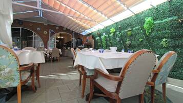 An indoor dining area with tables and chairs, a green wall with plants, and a tiled floor.