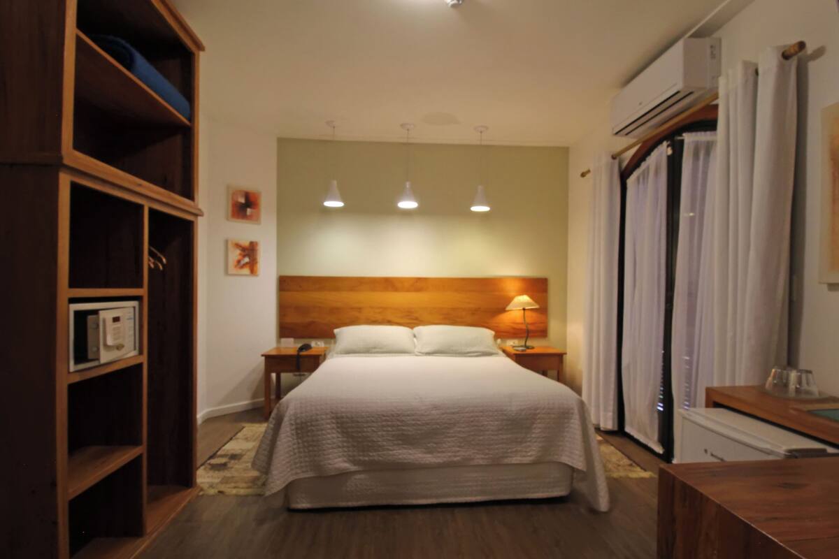 Une chambre à coucher avec une tête de lit en bois, un lit avec une literie blanche, une table de chevet avec une lampe, une armoire et une fenêtre avec des rideaux.